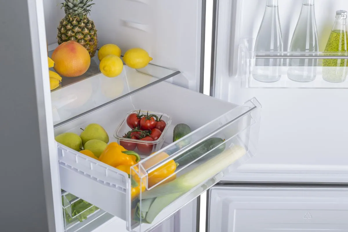 Белый холодильник с открытым ящиком для продуктов, наполненным фруктами и овощами