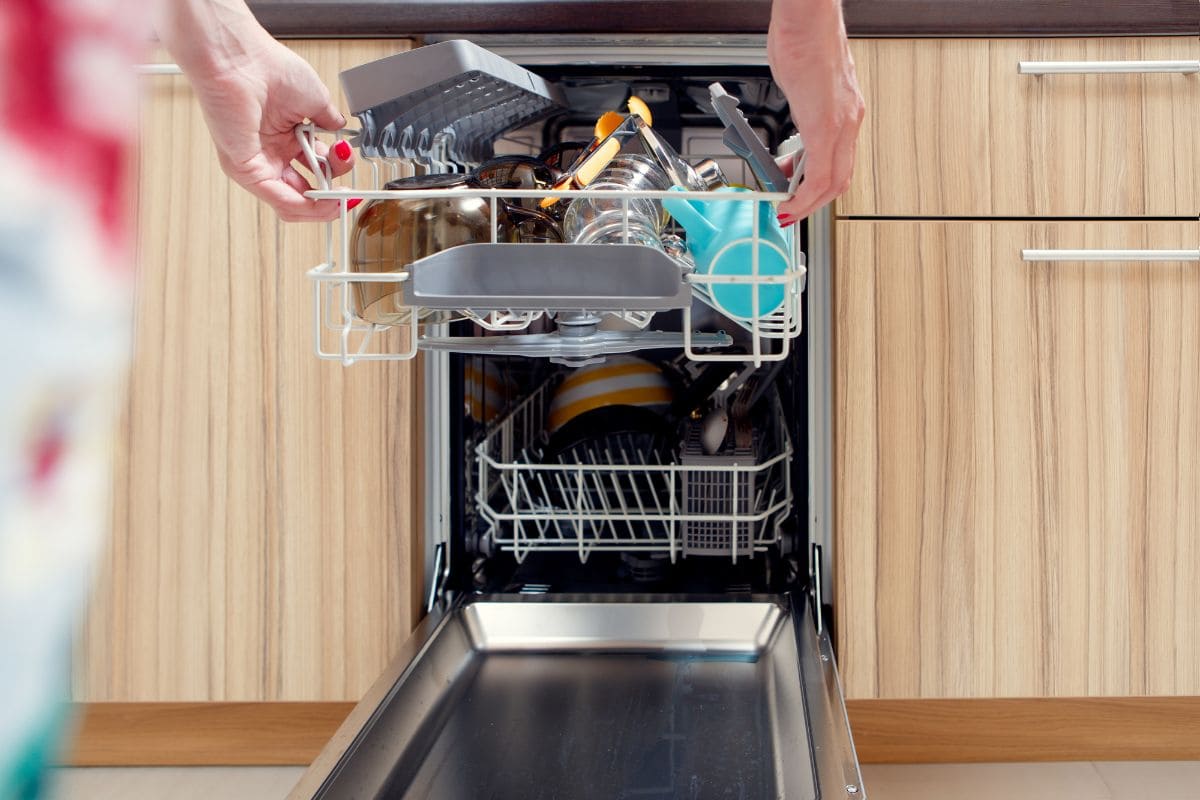 Женщина разгружает посудомоечную машину из нержавеющей стали, полную посуды с деревянными шкафами 
