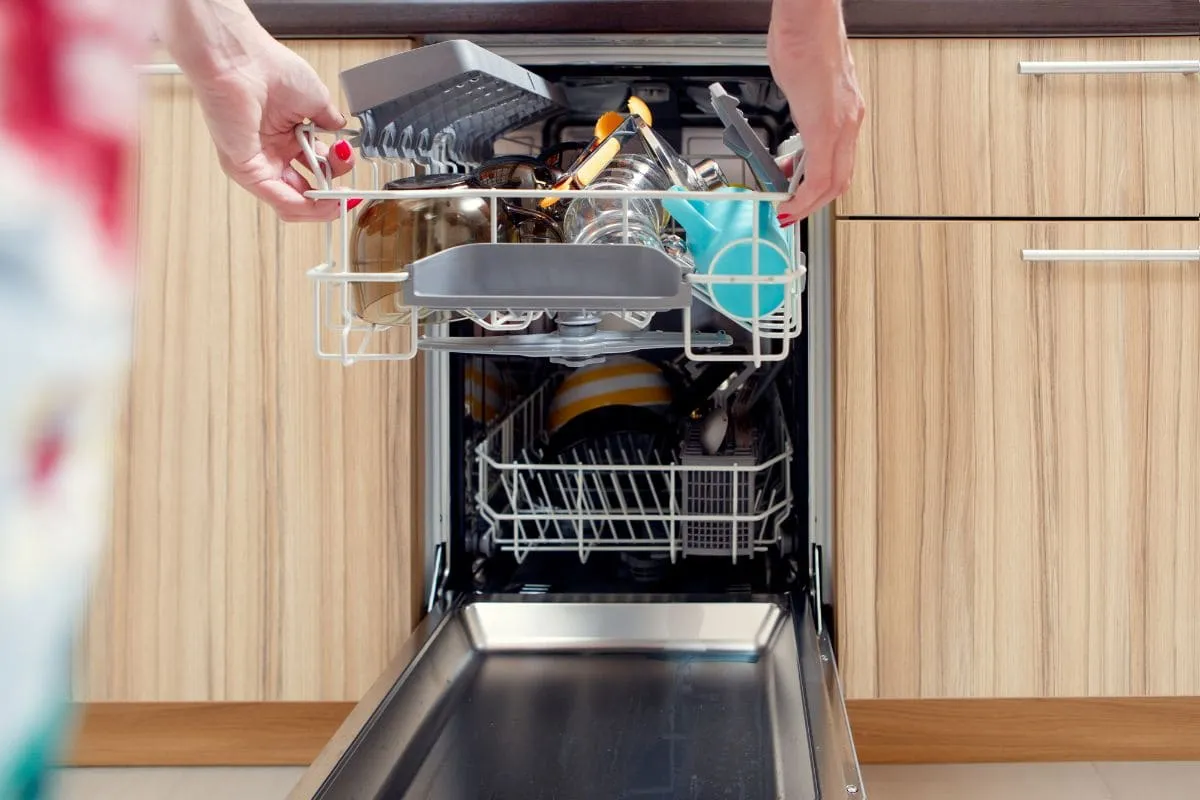Женщина разгружает посудомоечную машину из нержавеющей стали, полную посуды с деревянными шкафами 